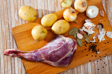Raw lamb leg on wooden cutting board, potatoes and condiments, ingredients for cooking