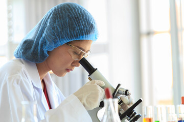 Asian female professional scientist in white lab coat safety glasses hygiene cap and rubber gloves using microscope zooming sample on laboratory working desk full of glass test tubes beaker and flask