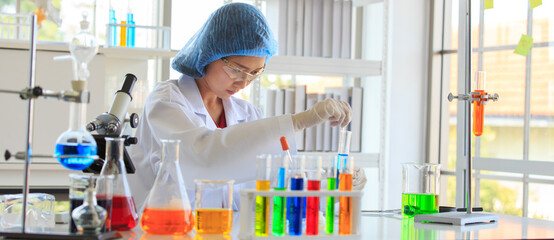 Asian female professional scientist in white lab coat safety glasses hygiene cap and rubber gloves using glass stick stirring liquid sample in test tube on laboratory analysis research working desk