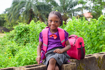 An African girl child in school uniform and with a backpack, sitting outside on a low fence