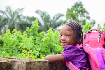 An African girl child in school uniform and with a backpack, resting her hands on a low fence