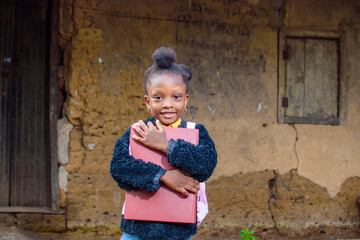A happy little African girl child or student with pink school bag, holding and hugging her books outside a village mud house - obrazy, fototapety, plakaty