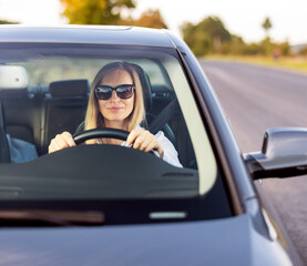 Confident mature lady with blond hair sitting inside modern car and keeping hands on steering wheel. Female driver wearing sunglasses and formal blue shirt.