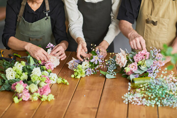 Close up of three florists arraigning flower compositions on wooden table in cozy workshop, copy...