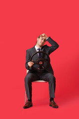 Tired young businessman with steering wheel sitting on chair against color background