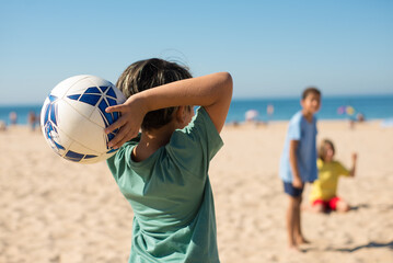 Rear view of preteen boy throwing ball on beach. Boy playing football at seaside. Youth sport,...