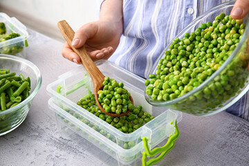 Woman putting frozen peas in plastic container at table in kitchen, closeup