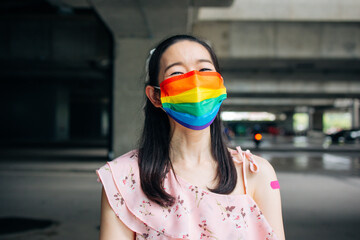 Portrait of smiling young Asian woman with covid-19 vaccination bandage standing wearing rainbow...