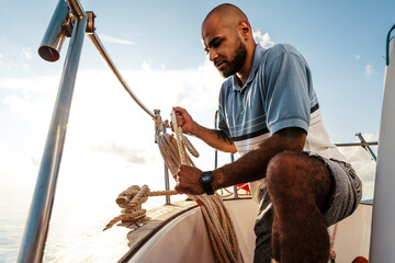 Young african american sailor tying ropes on sailboat in the sea on sunset