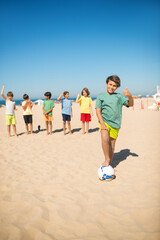 Happy preteen boy playing football with friends on beach. Kid standing with ball on sand and showing thumbs up gesture. Summer vacation, friendship concept