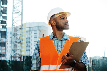 Young man engineer in workwear standing in construction site with clipboard