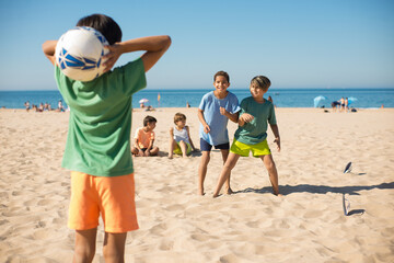 Cheerful boy friends playing football on beach. Preteen boy standing on sand and throwing ball to...