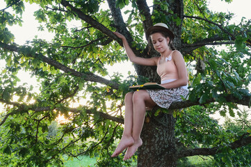 beautiful young girl in summer dress and hat is sitting on tree, on branch of large oak in sunset rays and reading book. Idea of summer vacation in nature