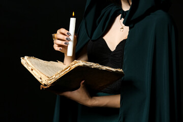 Young witch with spell book and candle on dark background