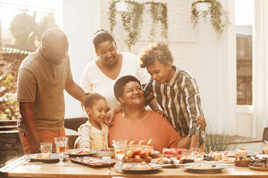 Portrait of happy African-American grandmother celebrating Birthday with family during dinner outdoors, lit by sunlight - Powered by Adobe