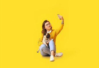 Young woman with cute pug dog taking selfie on yellow background