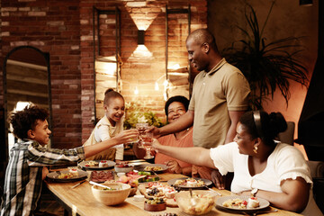 Warm toned portrait of happy African-American family clinking glasses and toasting while enjoying dinner together at evening - Powered by Adobe