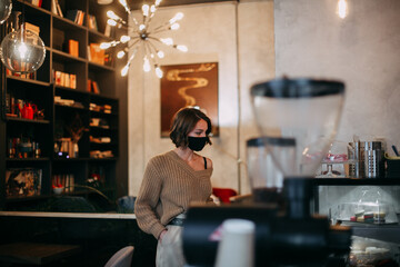 A young brunette girl in a brown sweater stands in a cafe at the showcase in a mask