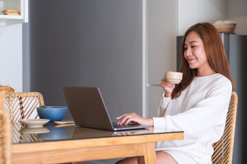 A young woman freelancer drinking coffee while using laptop computer for working or studying online at home