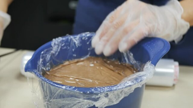 The female hands of a pastry chef in pink gloves are covering liquid chocolate in a bowl with a stretch wrapper for storage until cooking. Close-up, DOF, slow motion