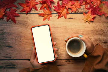 Woman holding smart phone and coffee cup wooden table with autumn maple leaves.Autumn season...
