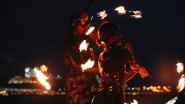 Fire show performance - man and woman in a shiny clothes playing with fire torches on the beach at night