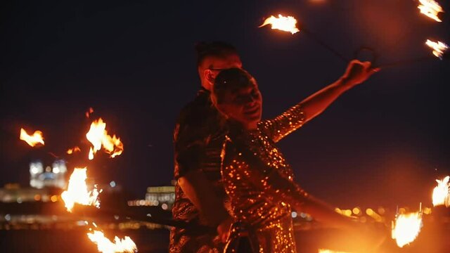 Fire show performance - smiling man and woman in a shiny clothes dancing with fire torches on the night beach