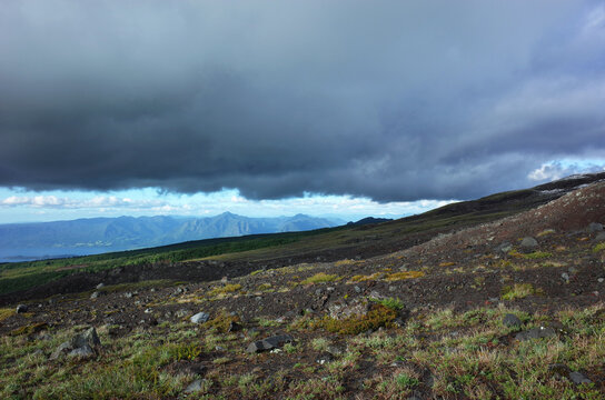 Dark slope of Villarrica volcano with sparse vegetation, low dark clouds and view of sunny mountains in distance between sky and earth, Nature background with space for text, Chile