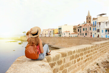 Beautiful young woman with hat sitting on wall looking at historic town of Alghero on Sardinia...