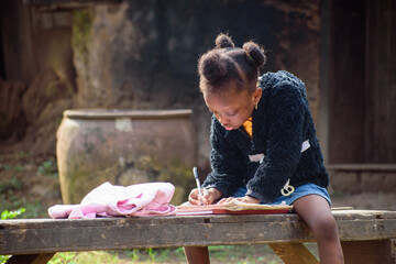 An African girl child or student sitting on a bench outside a village mud house, studying to attain...