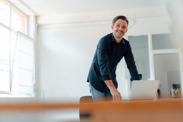 Smiling mature male professional with laptop at desk in board room at office