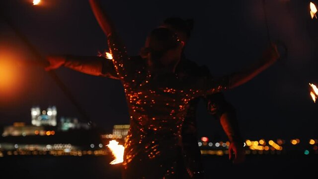 Fire show - smiling woman in a shiny dress and man behind her dancing with fire torches on the beach with a night city background