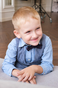 Vertical portrait of a five-year-old blonde boy in a shirt, vest and bow tie. Close-up of the child's face. The boy smiles and squints.