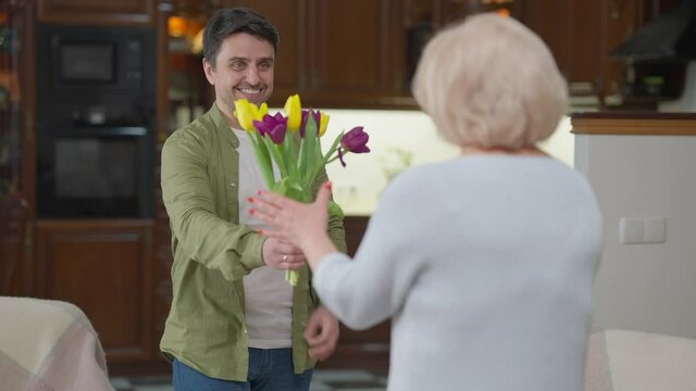 Happy handsome smiling adult man giving bouquet of yellow and purple tulips to senior woman indoors. Portrait of Caucasian son congratulating mother on birthday or Women's Day