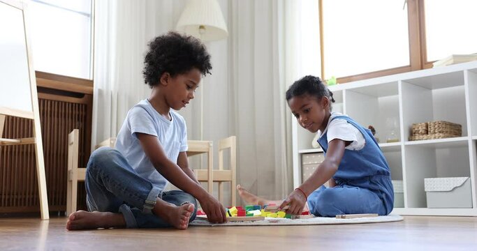 Amicable African beautiful siblings play together railroad at home, build wooden rail road parts, spend leisure time at modern playroom sit on warm floor. Children development game, friendship concept