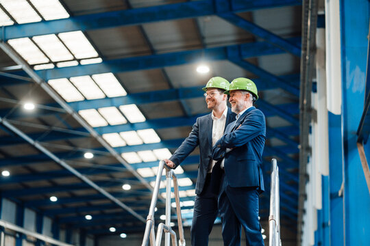 Smiling male business professionals wearing hardhat standing by railing in factory