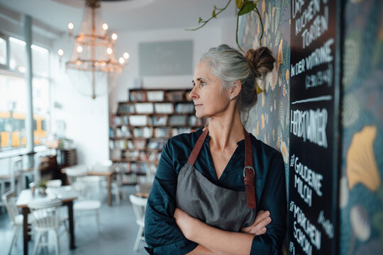 Female cafe owner with arms crossed leaning at blackboard in coffee shop