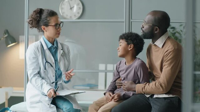 Cheerful female doctor sitting with little African American boy and his father on medical couch in clinic, talking with them and then giving high five to kid
