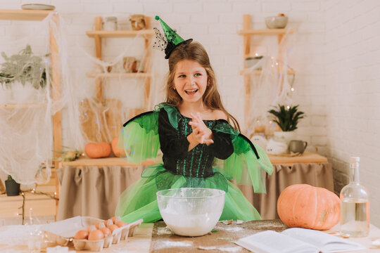 cute little girl in a green Halloween costume of a witch or fairy prepares a pumpkin pie, barm brack in a festive decorated kitchen. space for text. High quality photo