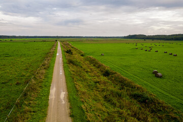 Aerial drone view shoot of rural dirty road leading through green farm lawn with straw bales and maize or rye plowed field and autumn forest. Agricultural parcels. Harvesting arable land, crop season