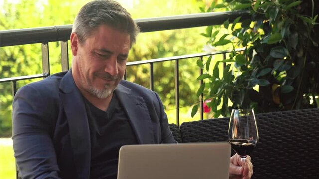 Professional wine expert reviewing red wine online using laptop computer at home on terrace. Online training, shopping, ordering wine