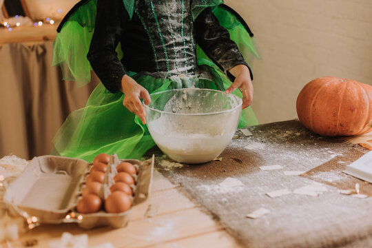 cute little girl in a green Halloween costume of a witch or fairy prepares a pumpkin pie, barm brack in a festive decorated kitchen. space for text. High quality photo