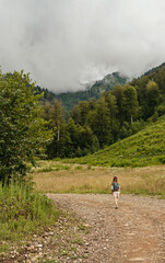 Rear view of young blonde woman with backpack walking uphill along trail on background of mountain...