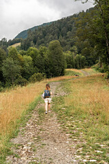 Naklejka na ściany i meble Rear view of young blonde woman with backpack walking uphill along trail on background of mountain covered with forest, active hiking eco tourism