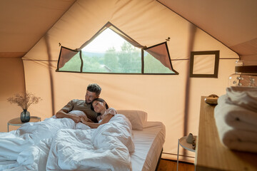 Affectionate couple looking at scenery around their glamping tent while lying in bed after sleep