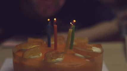 Close up with blurred background of blowing out candle fire on orange cake for birthday party with unrecognised person shows happy, joyful and memorable moment at home with family celebration.