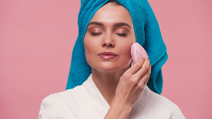 young woman with closed eyes using silicone facial cleanser isolated on pink