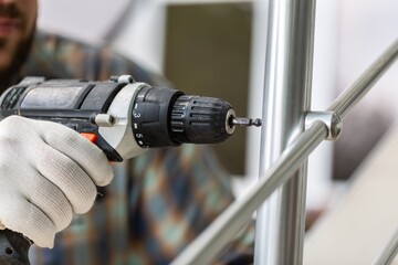 A worker in gloves repairs a metal railing of a staircase using a screwdriver and a drill