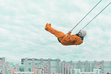 Astronaut wearing orange space suit and space helmet flying on a swing against cloudy sky above the...
