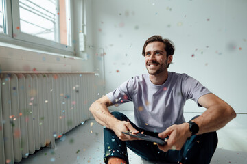 Man with digital tablet looking at confetti while sitting on floor